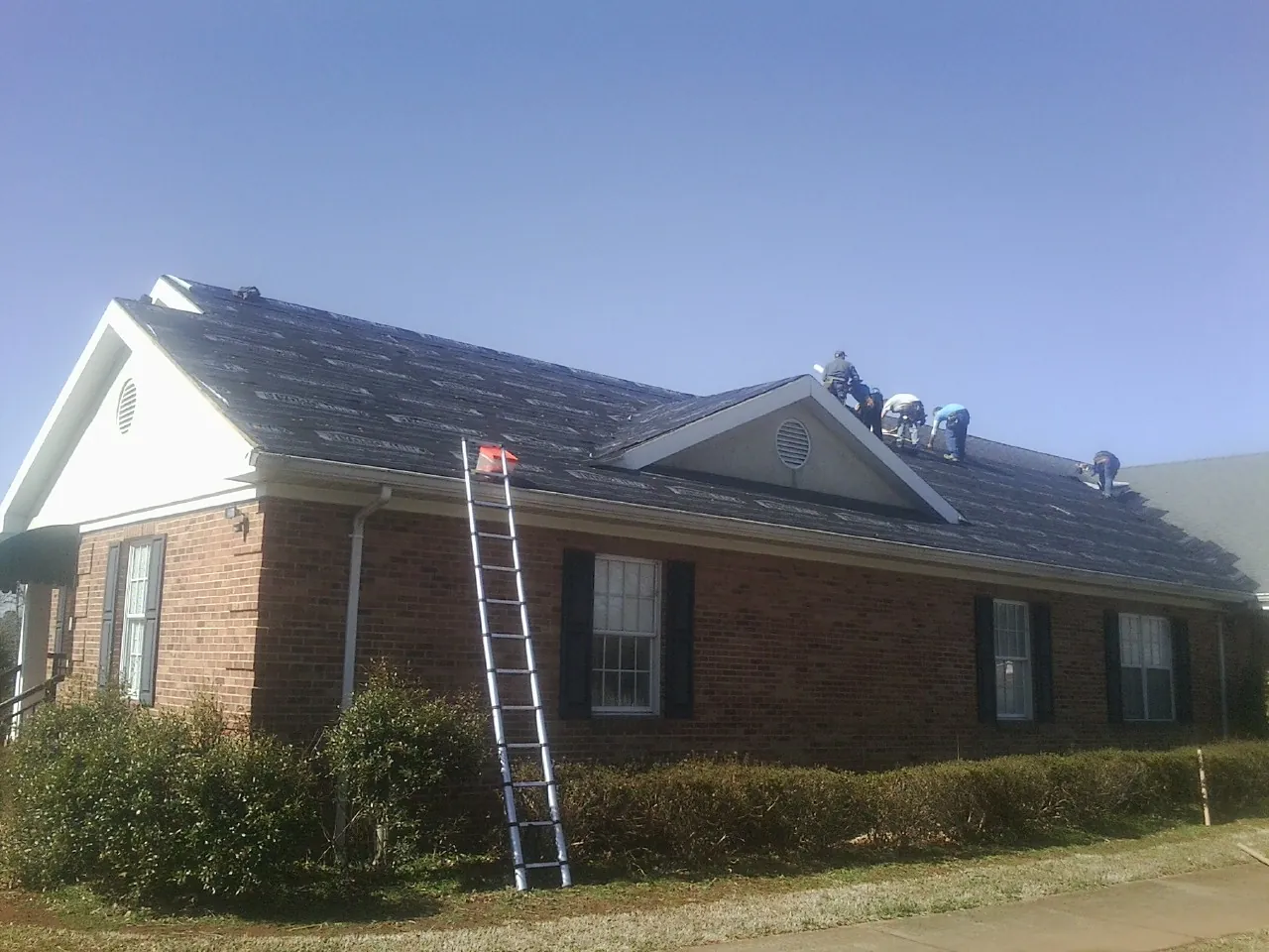 Crew tearing shingles off a brick ranch roof with a ladder set — Emergency Roof Repair work by our crew in Merry Hill